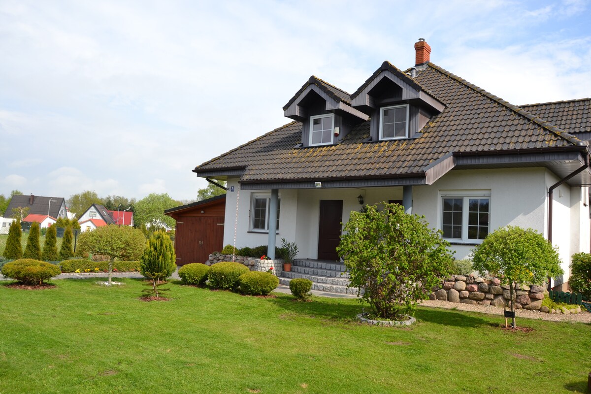 A two-story house with a sloped roof and dormer windows is set in a well-maintained garden. Lush green grass and neatly trimmed shrubs surround the entrance, which features stone steps leading up to the front door. A wooden storage shed is visible to the left.