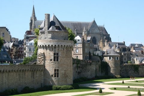 A historic castle stands prominently, featuring robust stone walls and round towers. Surrounding greenery complements the architecture. A visible church with a tall steeple is situated in the background, highlighting the blend of medieval structures and landscaped gardens.