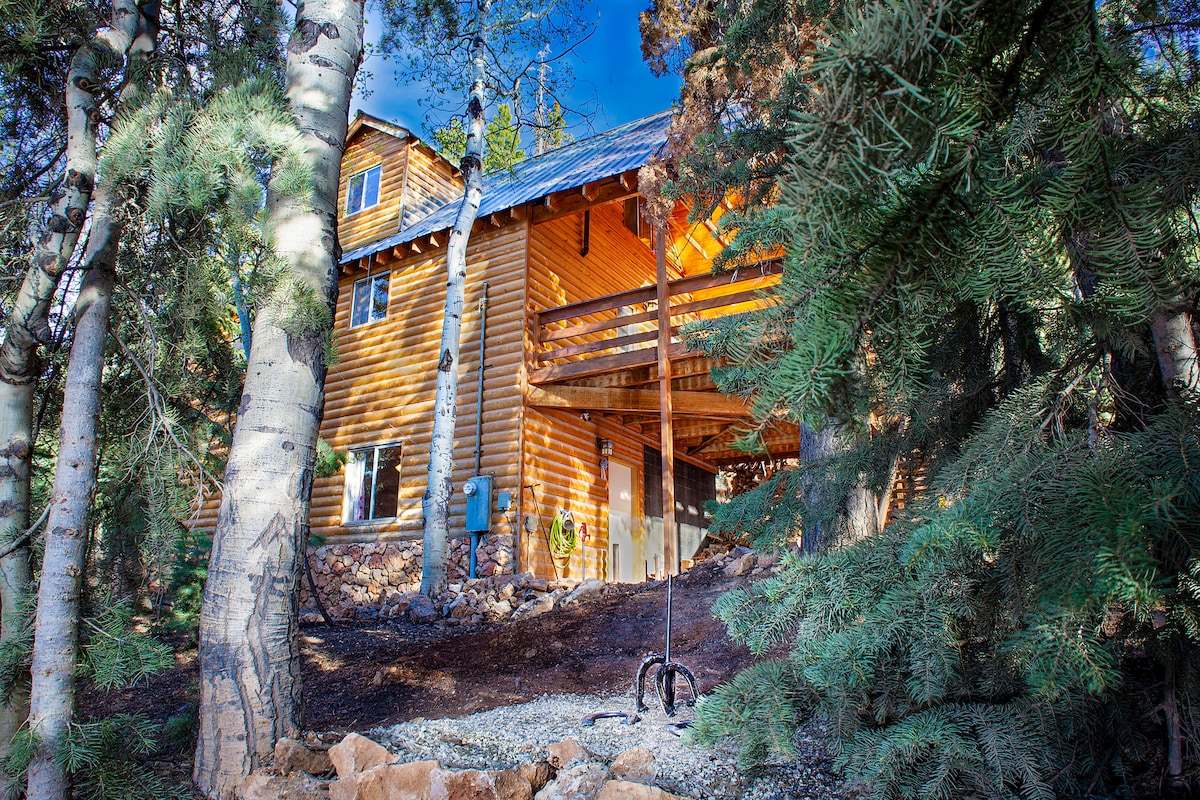 The exterior of a three-story cabin is surrounded by lush trees and natural landscaping. Large windows and wooden balconies are visible, allowing light to enter. The entrance is elevated and features a small staircase leading to the front porch, blending harmoniously with the natural setting.