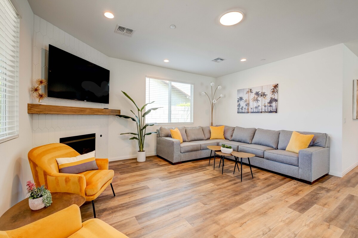 A spacious living room features a large sectional sofa with bright accent pillows. A yellow armchair adds a pop of color. A wall-mounted television is positioned above a wooden mantel, complemented by natural light from two windows. A simple coffee table sits at the center.