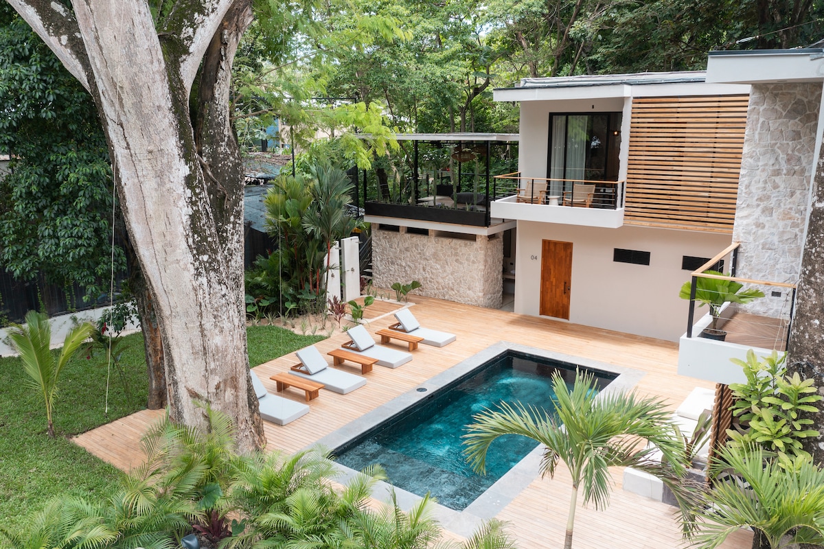 A modern outdoor area features a rectangular swimming pool surrounded by wooden decking. Four lounge chairs are positioned near the pool, with tropical plants and trees providing natural greenery. A spacious balcony with seating overlooks the pool, enhancing the tranquil atmosphere of the space.