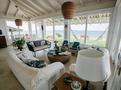 A light-filled living area features large glass windows facing the ocean, encouraging a connection with nature. Comfortable seating arrangements include a white sofa and lounge chairs. A large woven basket table sits in the center, complemented by decorative elements. A serene atmosphere is created by the open layout.