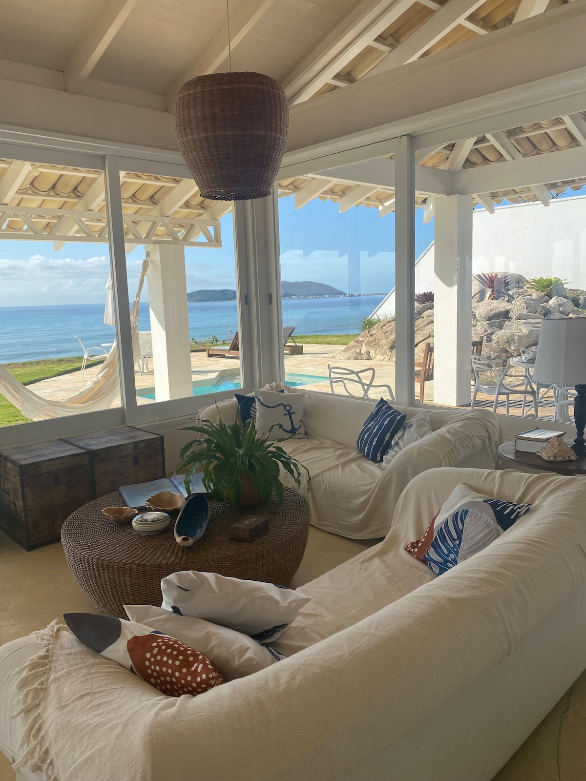 A spacious living area features large glass windows that provide views of the ocean and the outdoor seating area. A comfortable white sofa is complemented by decorative pillows, while a round coffee table with a plant adds a touch of nature. A hammock is visible in the background.