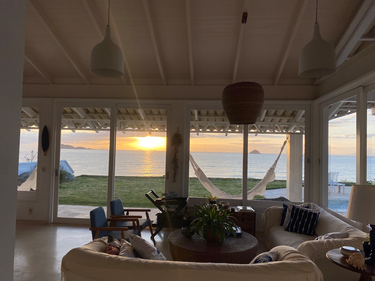 A spacious living area features large glass doors that frame a sunset over the ocean. Comfortable seating surrounds a round coffee table, while hammocks are seen outside on the lawn. Soft lighting from pendant fixtures enhances the peaceful ambiance.