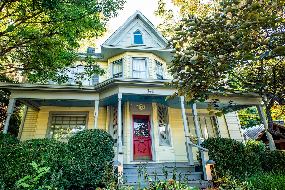 A historic Victorian home with a light yellow exterior is framed by lush green shrubs and trees. The inviting front porch features steps leading to a red front door, while large windows showcase the home's charm.