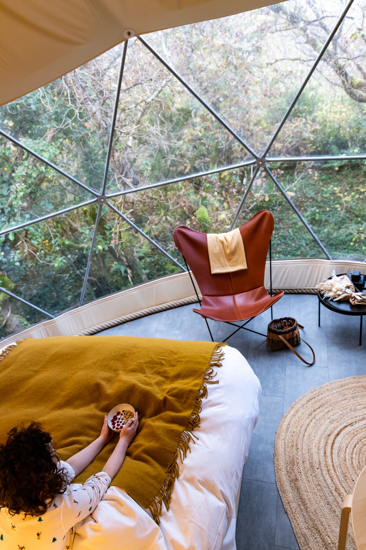 A spacious bed is dressed with a mustard-colored blanket. A person is seated on the edge, holding a bowl. A modern chair, designed in a butterfly shape, is placed near a small round table beside a woven rug. Natural light filters through the geodesic glass structure.