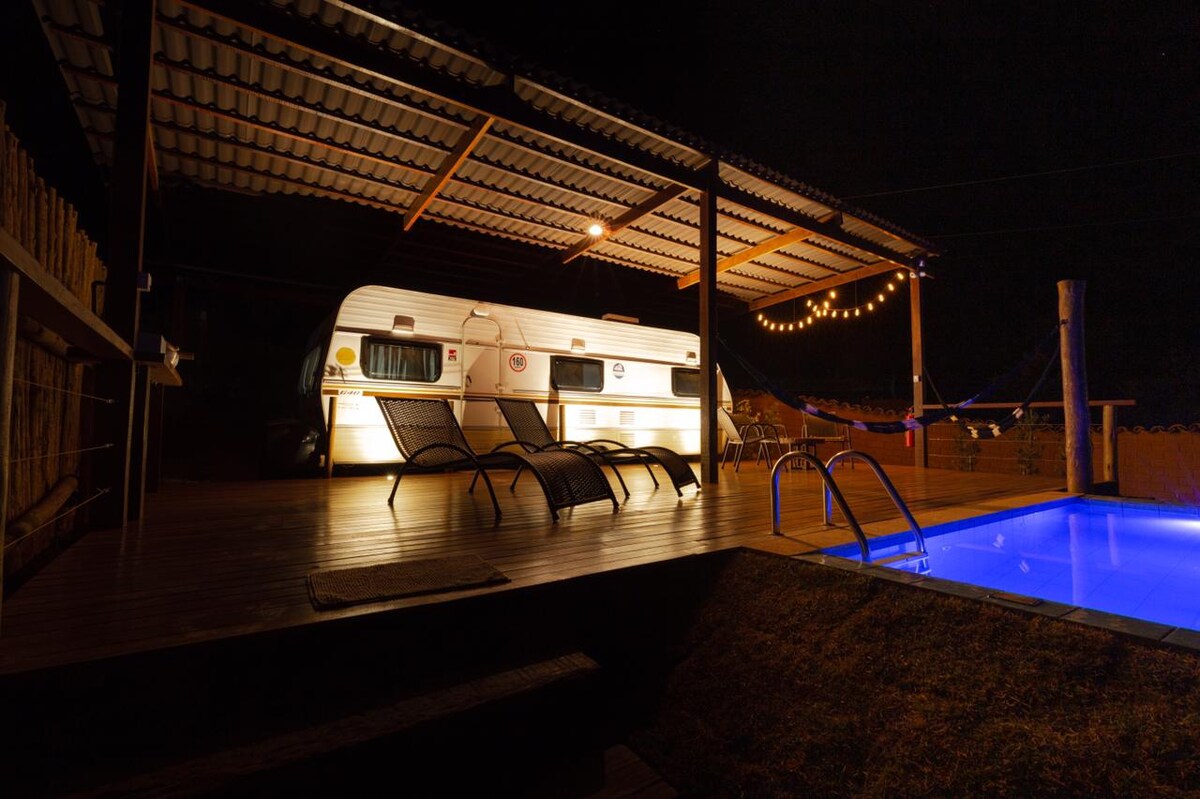 A vintage trailer is positioned under a covered patio, illuminated softly by string lights. Two lounge chairs are arranged beside a swimming pool with a gentle blue glow, creating a serene evening atmosphere in a tranquil outdoor setting.