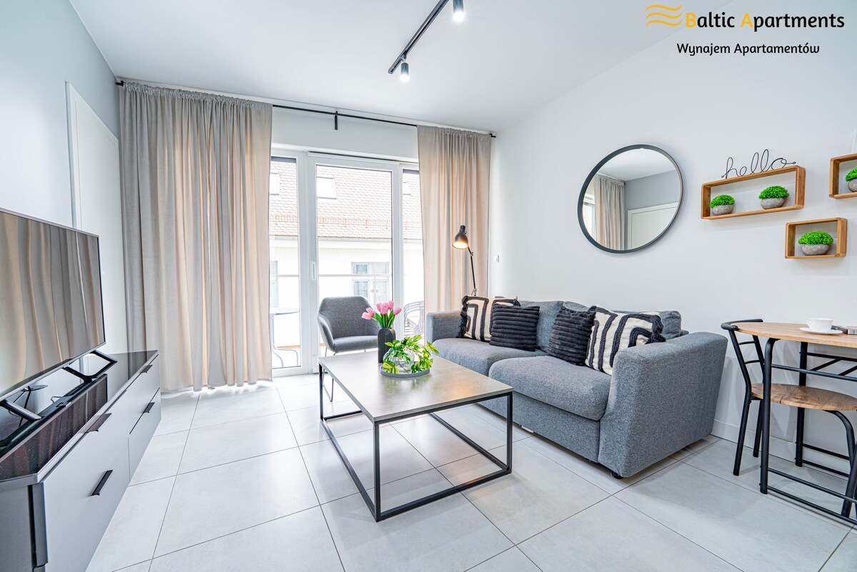 A modern living area features a comfortable gray sofa adorned with decorative pillows. A round mirror hangs above a sleek coffee table, complemented by a small plant. Natural light filters through sheer curtains, illuminating the space while a flat-screen television is positioned opposite the sofa.
