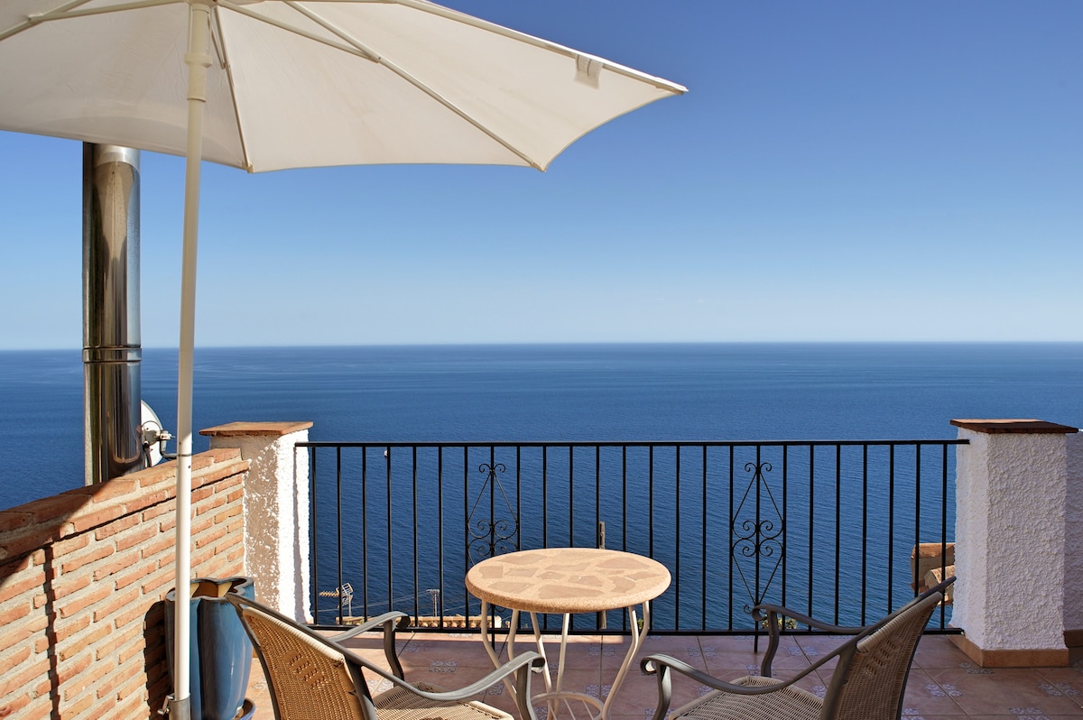 A terrace features a round table with two chairs, shaded by a large umbrella. A railing offers an unobstructed view of the expansive Mediterranean Sea, under a clear blue sky. The warm tones of the brick wall provide a homey touch to the open space.