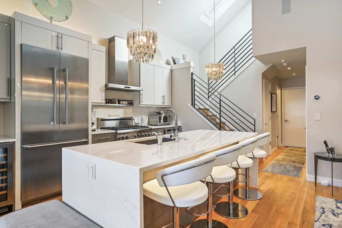 A modern kitchen features sleek stainless steel appliances and a large island with bar seating for four. Crystal chandeliers hang above, creating an elegant ambiance. A staircase leads to the upper level, while natural light streams in through a skylight, enhancing the airy atmosphere.