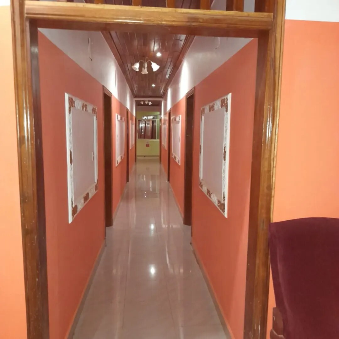 A hallway featuring a glossy tiled floor and warm orange walls. Framed artworks are displayed along the walls, leading to a well-lit area at the end. The ceiling features wooden beams, adding a touch of elegance to the space.