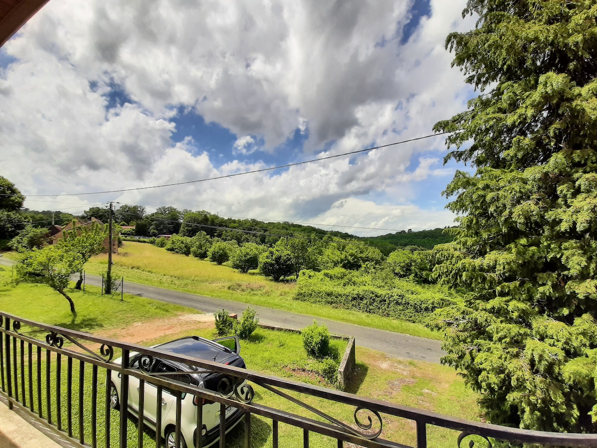 A view from the balcony captures a scenic landscape of lush greenery and distant hills under a partly cloudy sky. A quiet road runs through the property, with a parked vehicle visible on the grass, complementing the serene countryside setting.