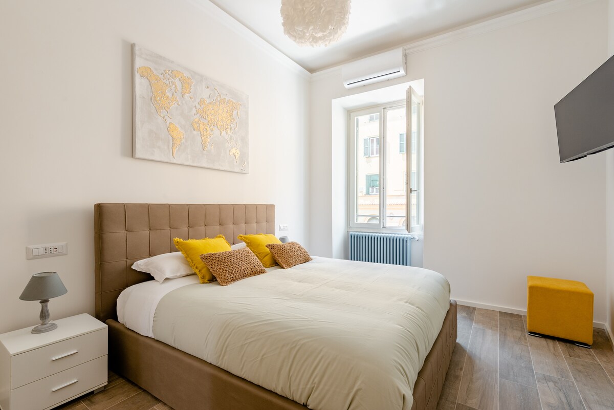 A bright bedroom features a spacious bed with neutral bedding and accent pillows. A large map artwork adorns the wall above the bed. A window allows natural light to enter, complemented by a small bedside lamp and a modern television mounted nearby.