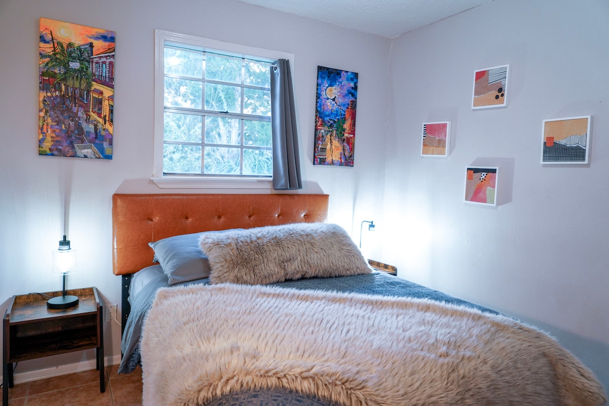 A cozy bedroom showcases a spacious bed with a plush blanket and decorative pillows. Colorful art pieces adorn the walls, and bedside lamps offer soft lighting. Natural light filters through a window, partially covered by a curtain, creating a warm and inviting space.