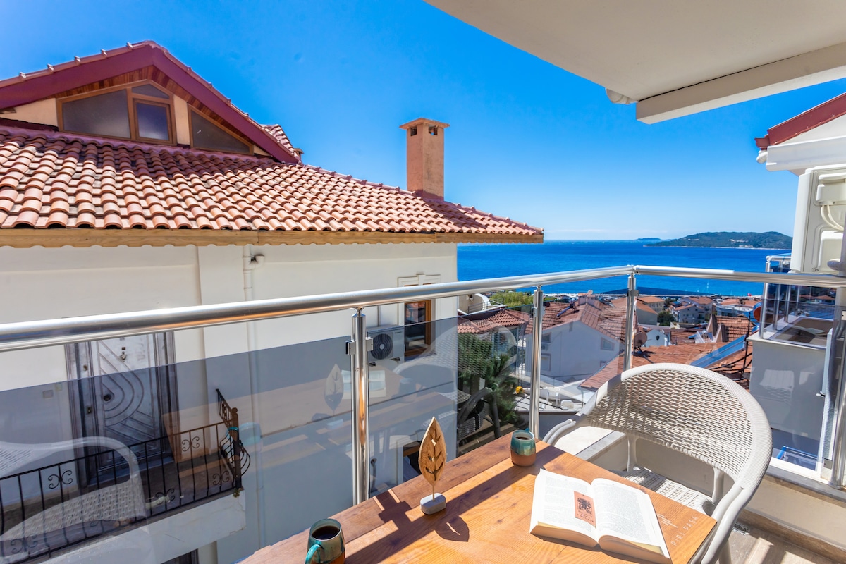 A balcony offers stunning views of the ocean and surrounding rooftops, featuring a wooden table with an open book and two tealight candles. A white wicker chair is positioned nearby, inviting relaxation. The clear blue sky complements the serene atmosphere.