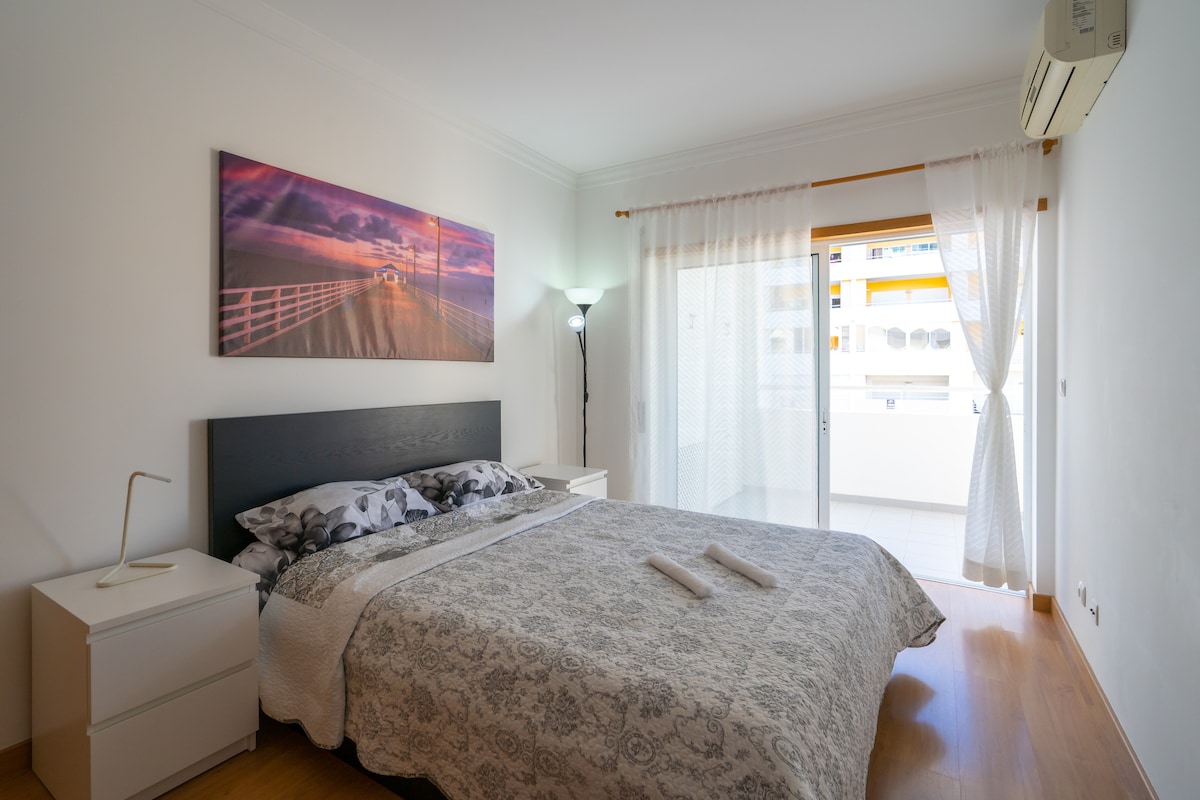 A bright bedroom features a spacious bed dressed in a light-colored coverlet. A large window allows natural light to flow in, complemented by sheer curtains. A bedside lamp and two nightstands create a minimalistic decor, while a piece of artwork adds a touch of color to the wall.