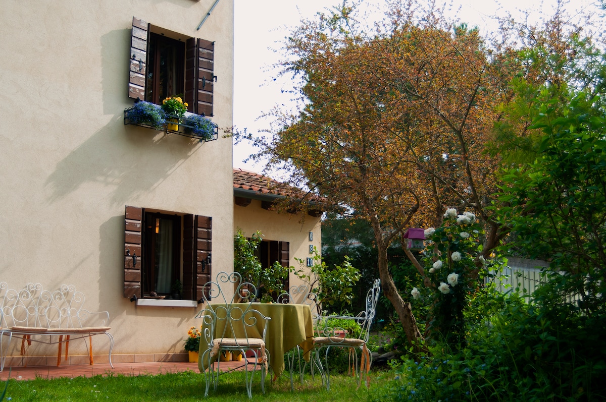 The exterior of a charming country house showcases brown shutters and a flower box filled with bright blooms. A small round table is set with chairs in a green garden, surrounded by lush greenery and flowering plants, creating a serene outdoor space.