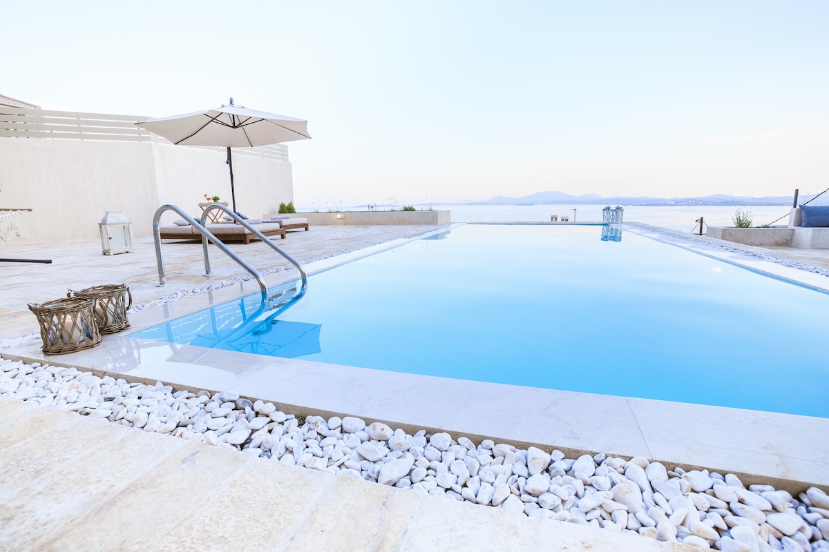 A private swimming pool is surrounded by a smooth stone deck and landscaped with decorative pebbles. An umbrella shades a lounge chair nearby, while a scenic sea view is visible in the background, enhancing the tranquil space.