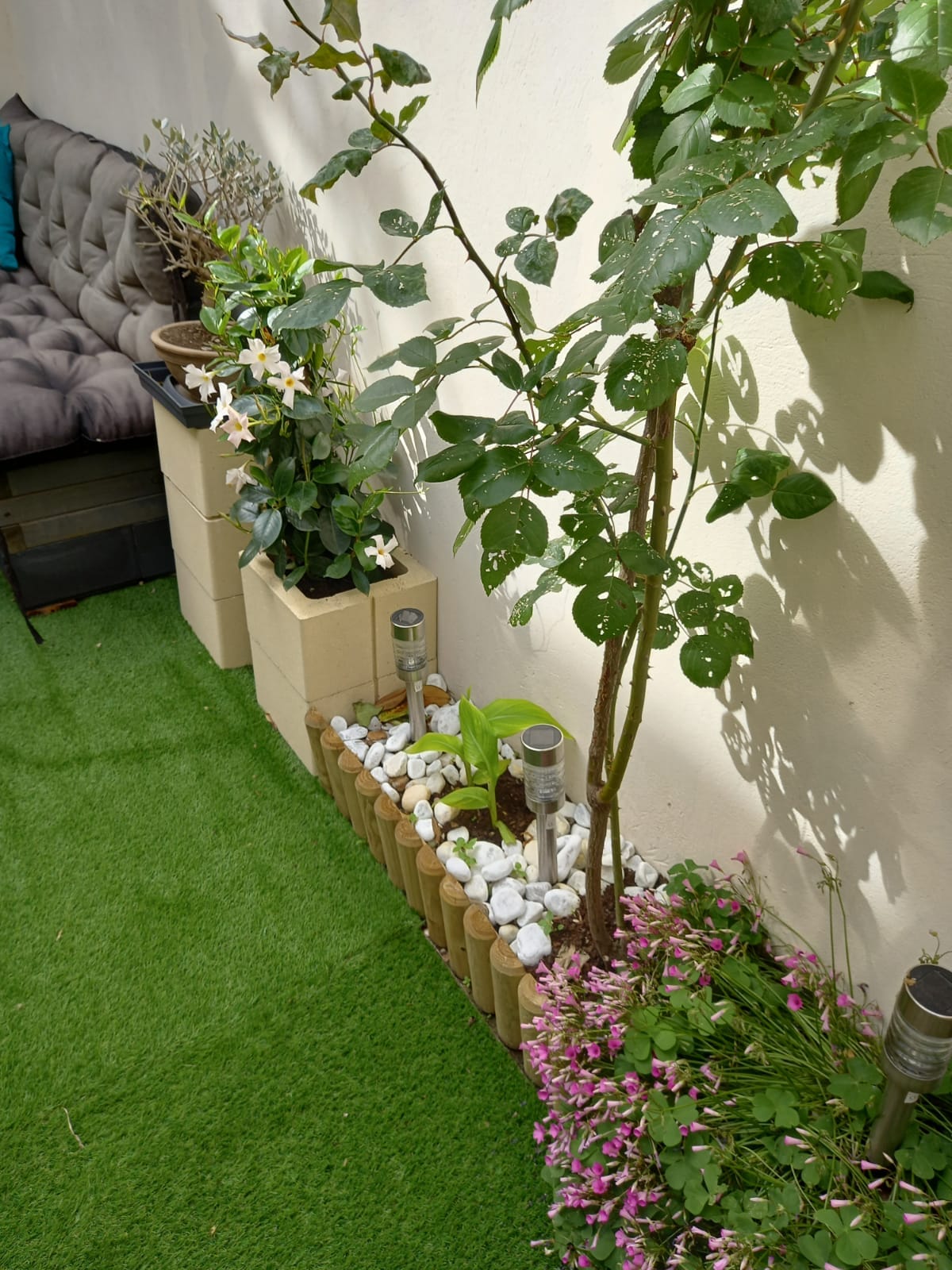 A serene outdoor space is presented, featuring lush green grass and decorative planters. Various plants, including flowering ones, are arranged alongside decorative stones and bamboo-like elements. Soft seating is also visible, providing a comfortable area for relaxation.