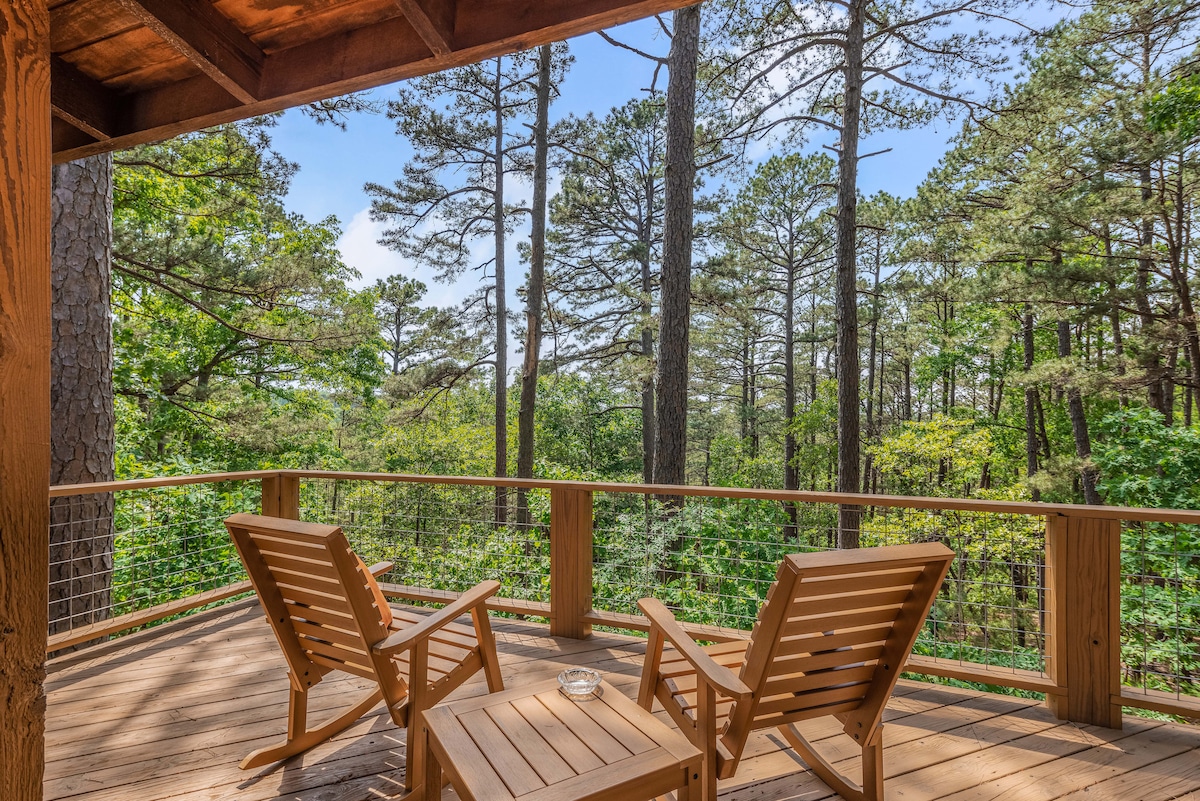 A private deck is furnished with two wooden chairs positioned around a small table, offering a serene view of the surrounding wooded landscape. Tall trees create a natural backdrop, allowing for a peaceful outdoor experience.
