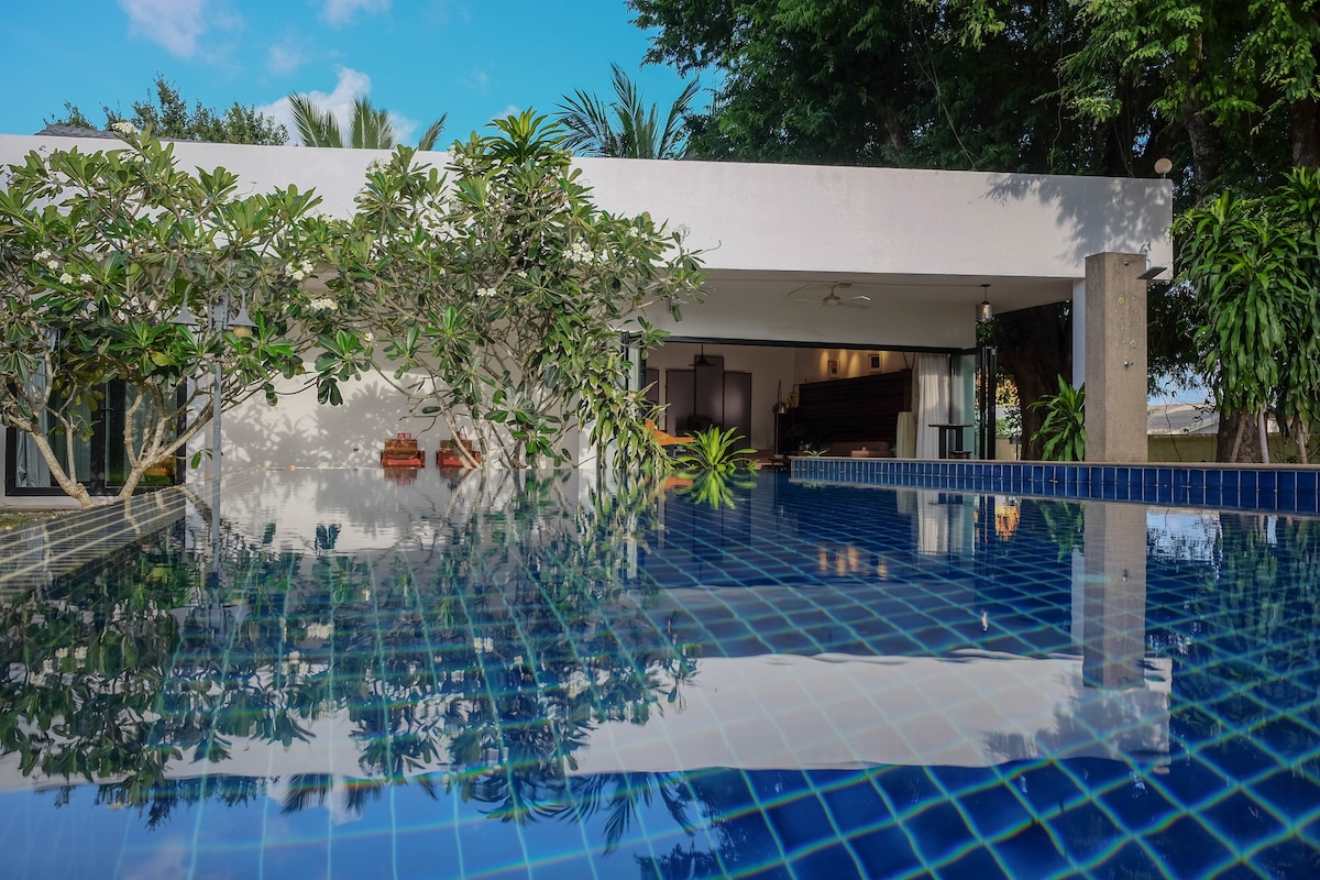 An outdoor saltwater pool reflects the vibrant blue sky and surrounding greenery. The modern architecture of the villa is visible, with large glass windows offering glimpses of the living spaces within. Lush plants frame the pool area, enhancing the natural setting.
