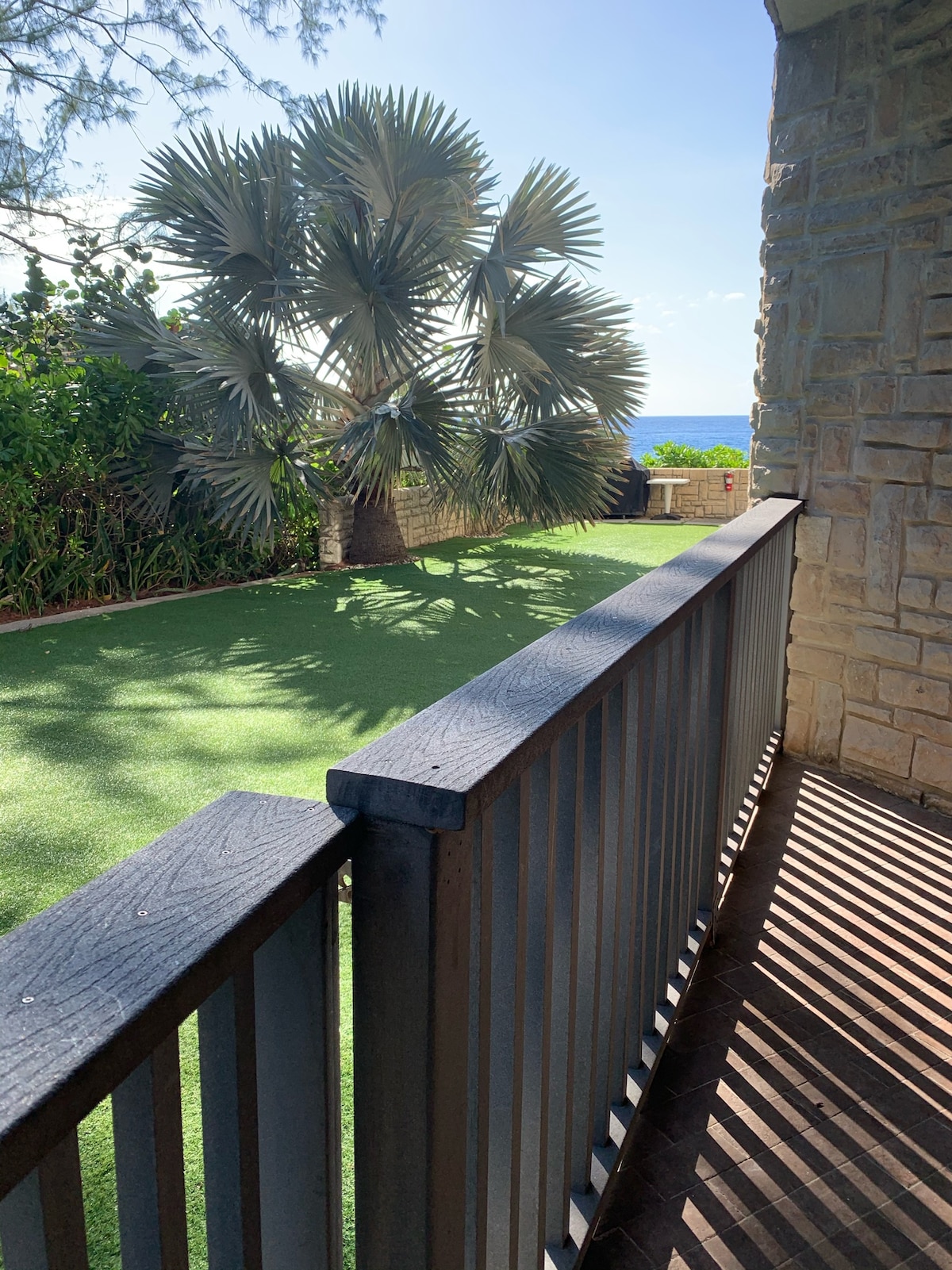 A view from a private patio showcases a well-maintained green space with a palm tree. The patio's railing casts interesting shadows on the floor, while the sun shines from a clear sky, enhancing the outdoor ambiance.