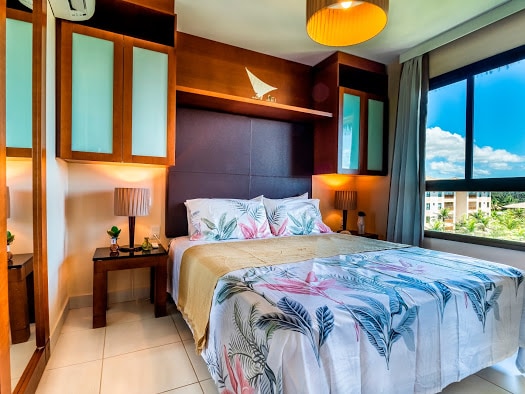 The bedroom features a spacious bed adorned with a floral-patterned blanket. Soft lighting from bedside lamps creates a relaxing ambiance. Large windows provide natural light and a view of greenery, while wooden cabinetry offers storage space.
