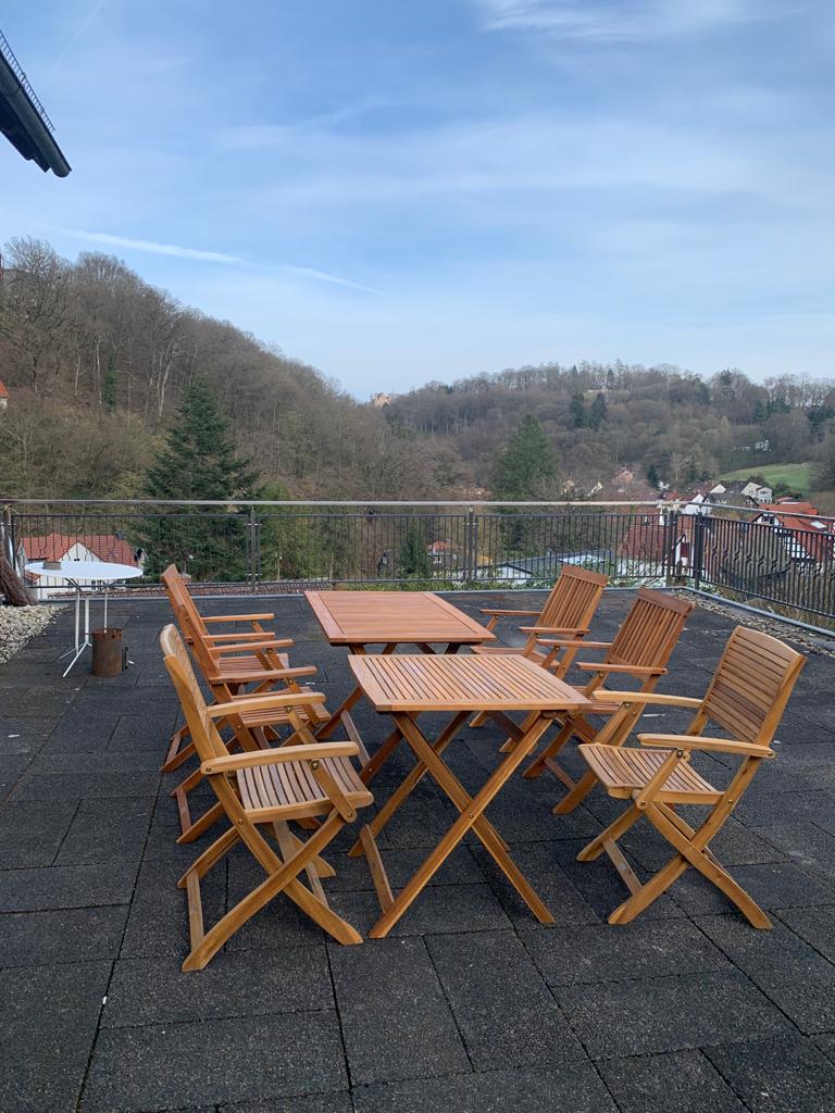A spacious terrace is outfitted with wooden dining furniture, including a large table surrounded by six matching chairs. The area is enclosed by a railing, offering scenic views of the surrounding green hills and nearby village.