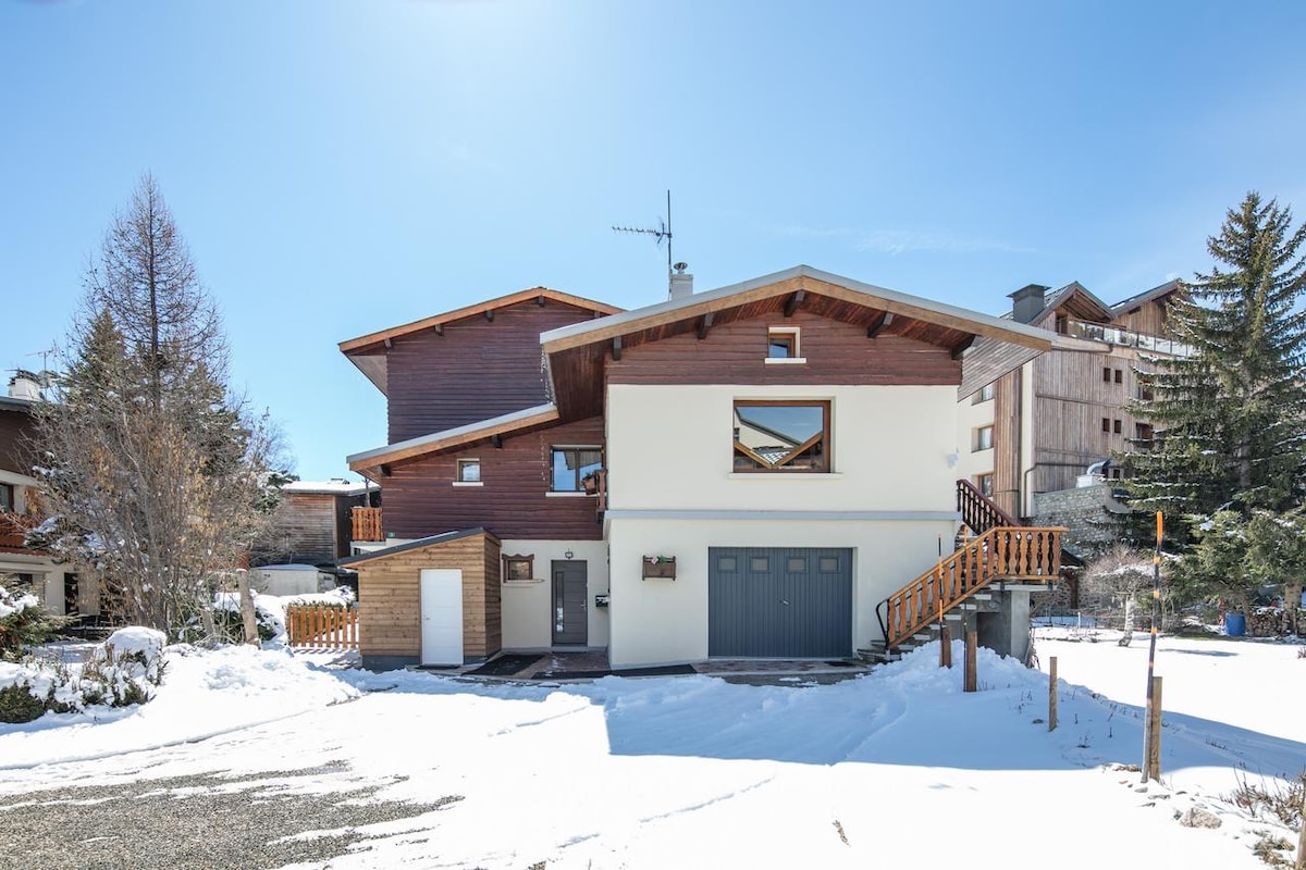 The exterior of a wooden chalet is displayed, featuring two stories with a blend of dark and light wood accents. Snow blankets the ground, with a well-maintained driveway leading to the front door. Surrounding trees and nearby buildings contribute to the winter scenery.