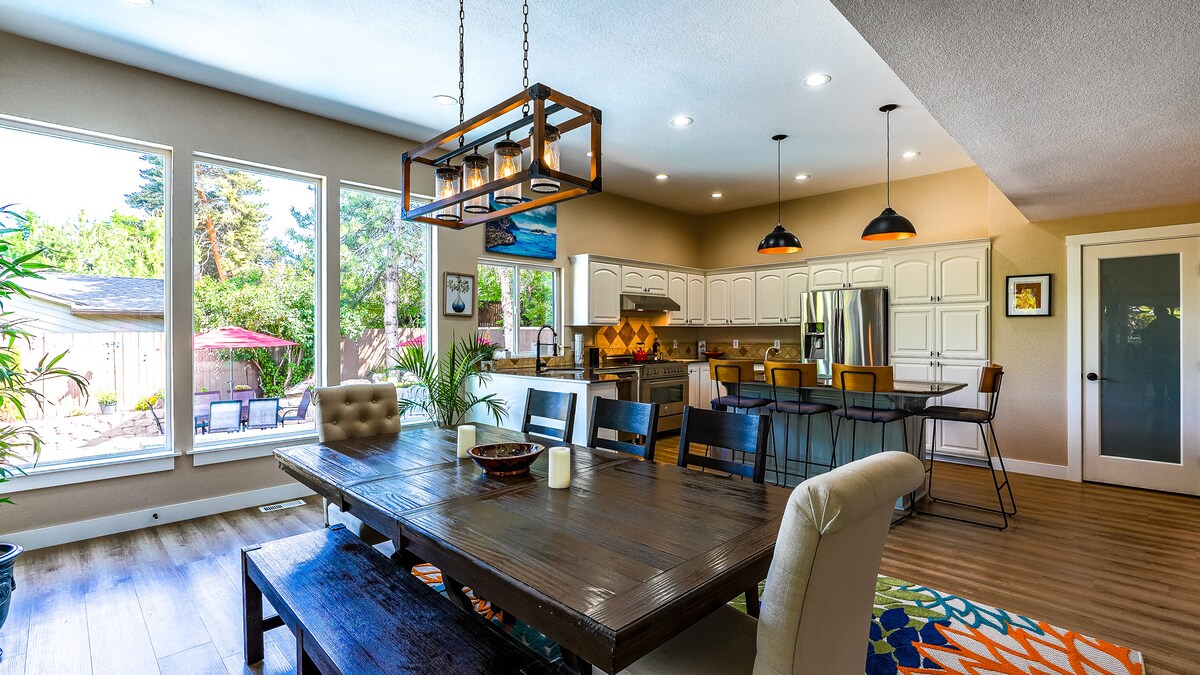 A spacious dining area features a large wooden table with comfortable seating for eight. Full-length windows flood the space with natural light, offering views of the backyard. The well-equipped kitchen is visible, with modern appliances and decorative pendant lights enhancing the inviting atmosphere.