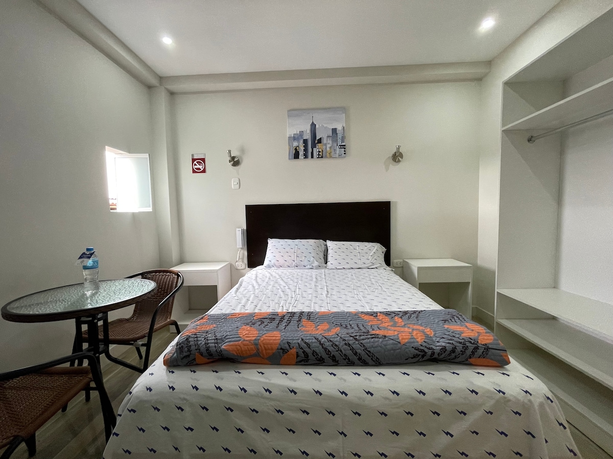 A cozy guest room features a spacious bed with patterned bedding and a soft throw blanket at the foot. A small round table with two wicker chairs creates a comfortable seating area. Natural light enters through a small window, illuminating the minimalistic decor.