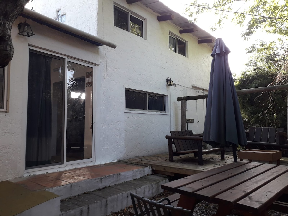 The exterior of the cabin is shown, featuring a wooden deck with a table and chairs. A large umbrella stands beside comfortable seating. Large glass doors provide access to the interior, while bright light filters through nearby trees.
