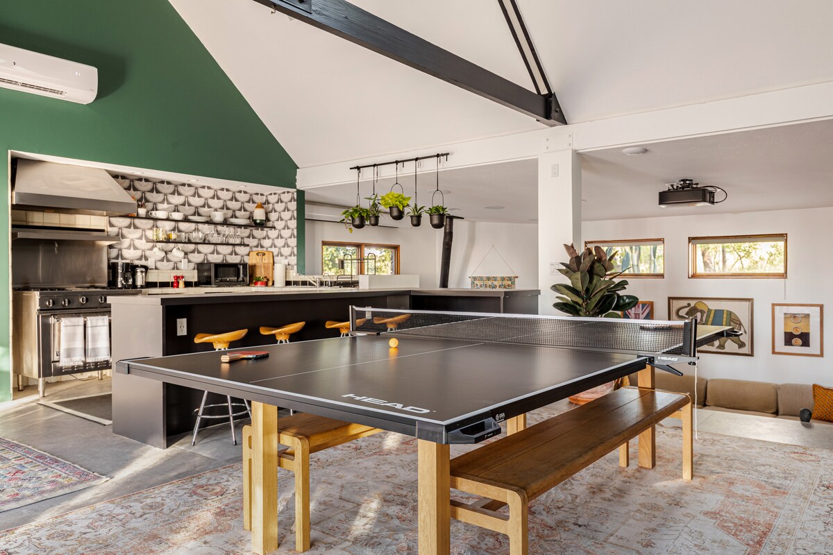 An open-concept living area is highlighted by a large table with a ping pong surface, surrounded by wooden benches. The kitchen area features modern appliances and a bar with three stools. Green walls and natural light enhance the inviting space.