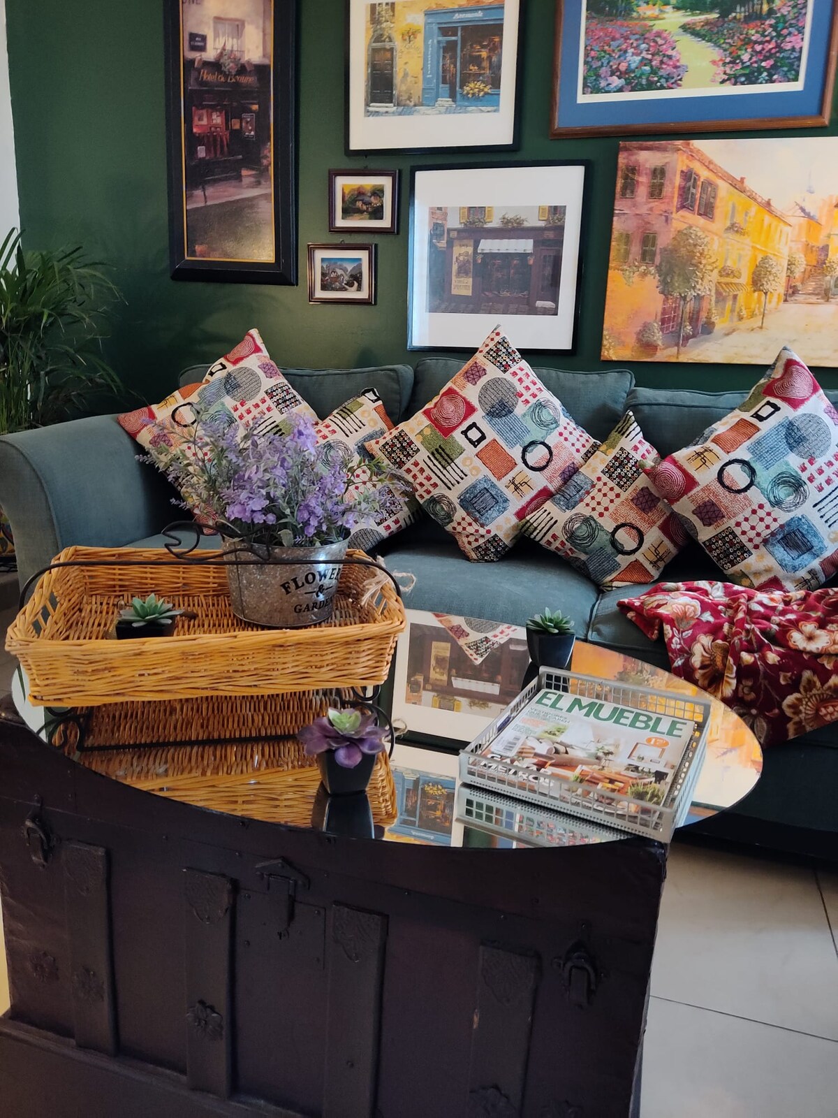 A cozy seating area features a green sofa adorned with patterned cushions, arranged against vibrant wall art. A round glass table sits in front, complemented by a woven basket and potted plants. Soft natural light enhances the inviting decor.