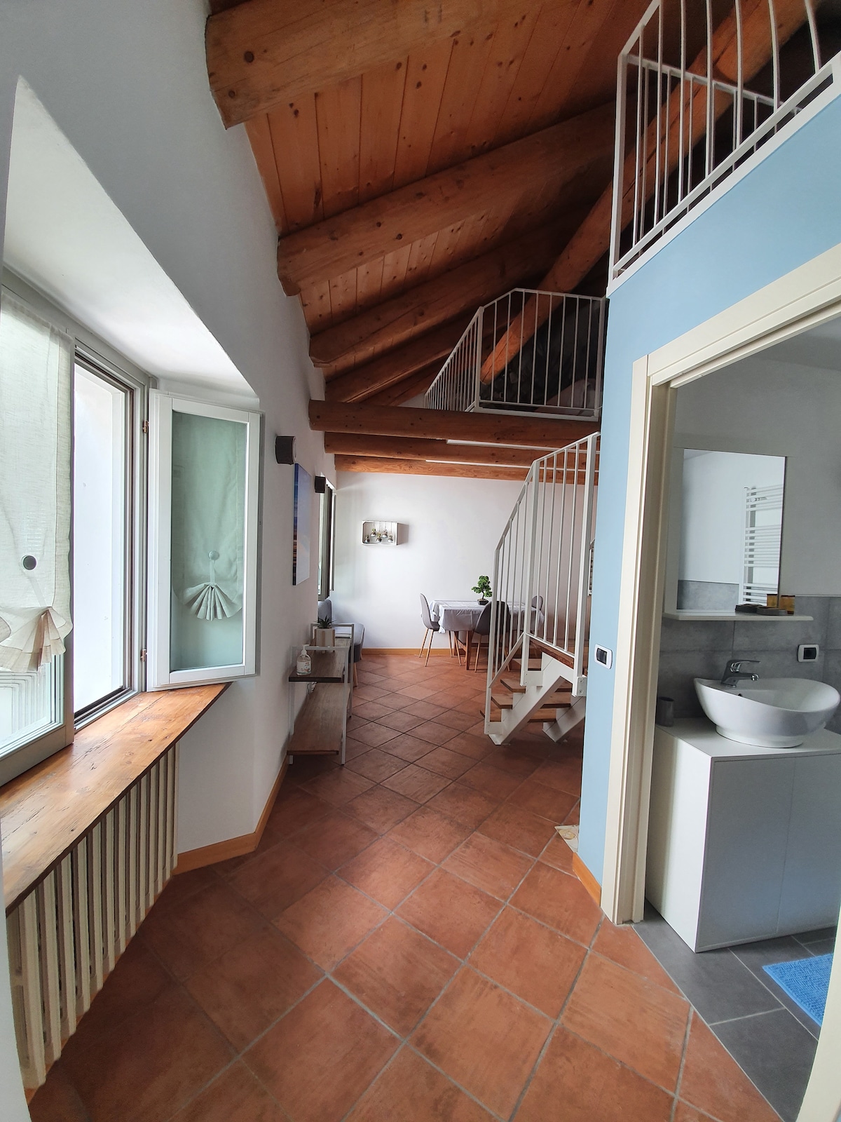 A bright interior space features sloped wooden ceilings and terracotta floor tiles. Large windows allow natural light to fill the area. A decorative table with chairs is visible, along with a spiral staircase leading to an upper level. The bathroom area is partially seen on the right.