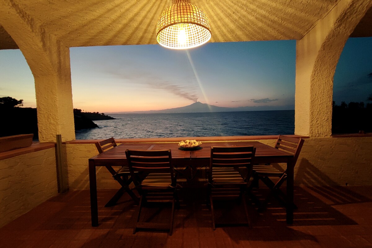 An outdoor dining area is presented under a large, woven pendant light. A wooden table with several chairs faces the sea, offering a stunning view of Mount Etna during sunset. The surrounding architecture includes arched openings, enhancing the scenic view.