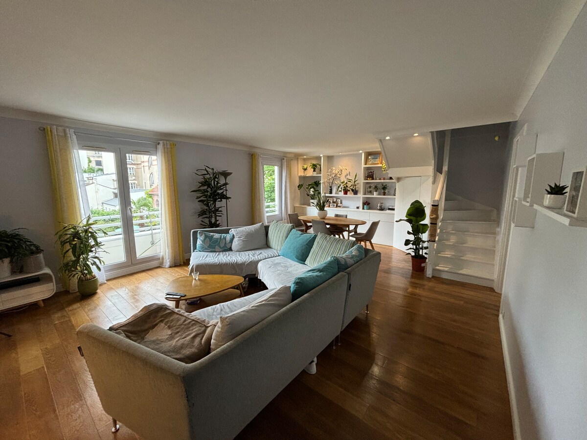 A spacious living area is enhanced by wooden flooring and large windows. A comfortable sectional sofa and a small coffee table occupy the center, while decorative plants add greenery. A staircase leads to the upper level, with an open kitchen and dining area visible in the background.