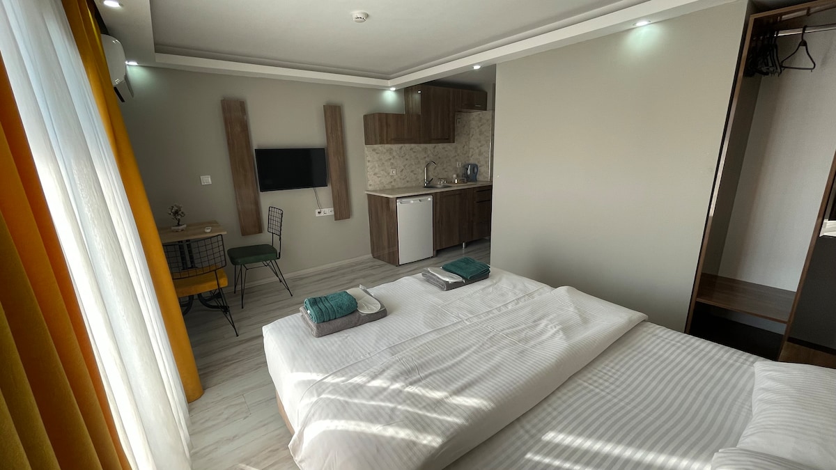 A well-lit room features two spacious beds with white linens, accompanied by complimentary towels. A kitchenette with modern cabinetry is visible, alongside a dining area with a small table and two chairs. A wall-mounted television is positioned for convenient viewing.