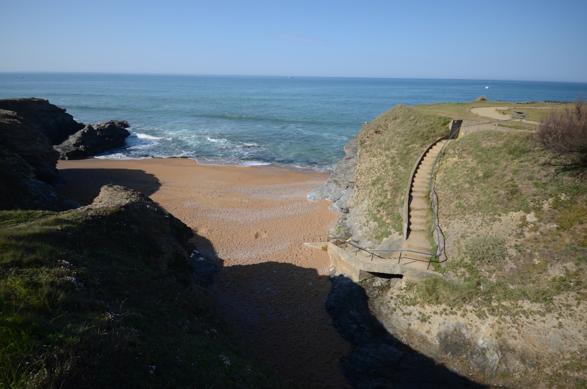 A tranquil beach scene features a sandy shore with gentle waves lapping at the water's edge. A staircase leads down from a grassy area to the beach, surrounded by rocky outcrops and sparse vegetation. Clear blue skies create a serene atmosphere.