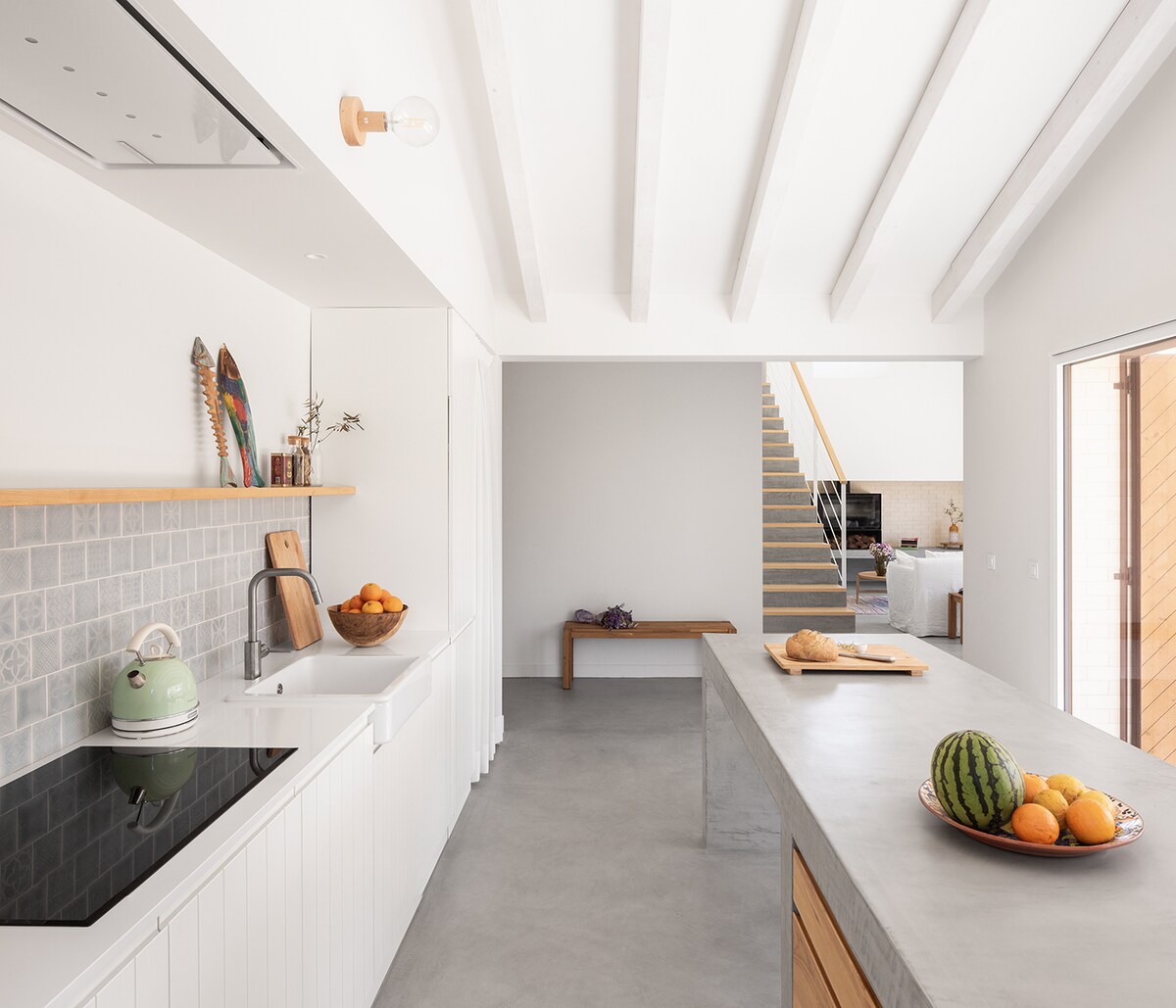 A modern kitchen area features a sleek countertop with an array of fresh fruits displayed on a wooden platter. A spacious sink and a stovetop are integrated into the clean, minimalist design. Wooden beams highlight the ceiling, while a staircase is visible in the background.