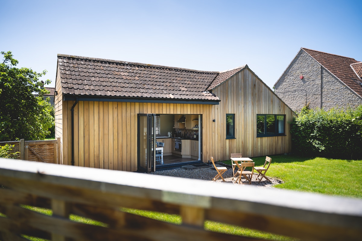 The exterior of the cottage is shown, featuring a wooden facade with large bifold doors that open to the garden. A small dining set is visible on the lawn, surrounded by greenery, while the roof is covered with traditional tiles. The surrounding structures are also partially seen.