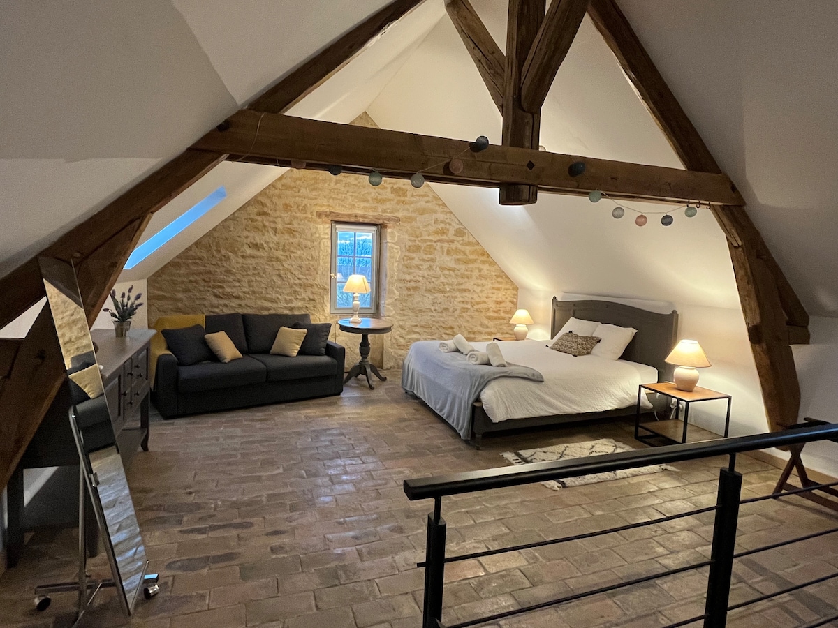 This spacious attic bedroom features exposed wooden beams and stone walls. A comfortable bed is positioned alongside a cozy sofa and a side table with a lamp. Natural light enters through a small window, creating a calming ambiance within the room.