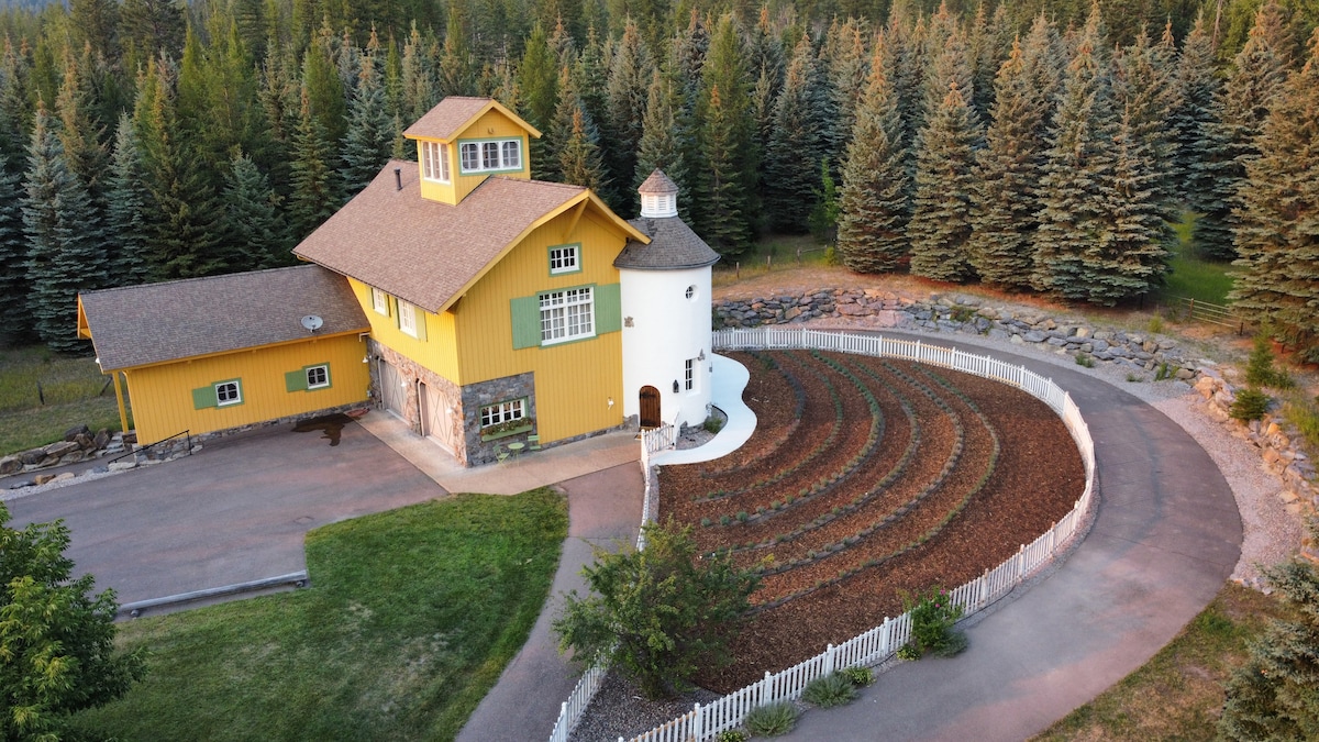 The property showcases a vibrant yellow exterior with a distinctive round turret. Surrounding greenery complements the structure, while a circular garden with neatly arranged raised beds is visible in the foreground, bordered by a white picket fence.