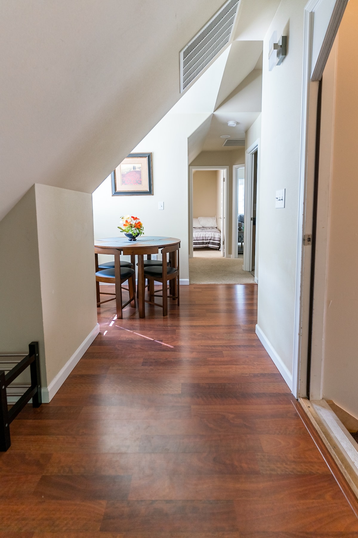 A hallway connects various spaces within the apartment. To the left, a small dining table with four chairs is set against a wall adorned with a framed print. Soft lighting highlights the warm wood flooring, leading toward a doorway opening to a bedroom.