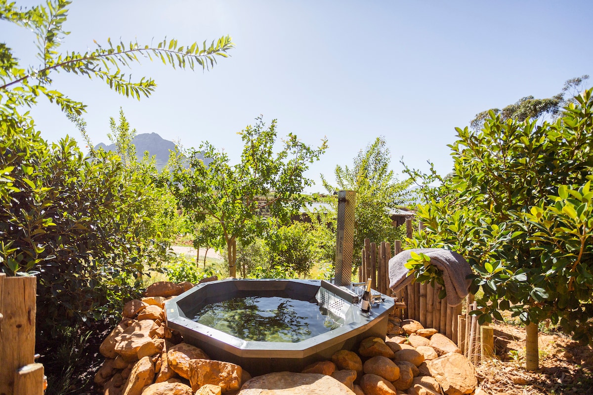 A wood-fired hot tub is nestled among lush greenery and natural stone, offering a serene spot for relaxation. The surroundings feature small trees and shrubs, creating a private atmosphere with views of the distant mountains under a clear blue sky.