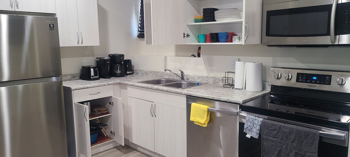 A modern kitchen is equipped with stainless steel appliances, including a refrigerator, dishwasher, and oven. White cabinetry provides ample storage, while a double sink is positioned under a window. Counter space features a variety of dishes and coffee machines, ensuring a functional cooking area.