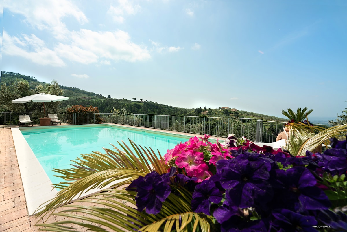 A serene swimming pool is framed by vibrant flowers in varying shades of purple and pink. Lush green hills can be seen in the background under a clear blue sky, creating a peaceful outdoor environment.