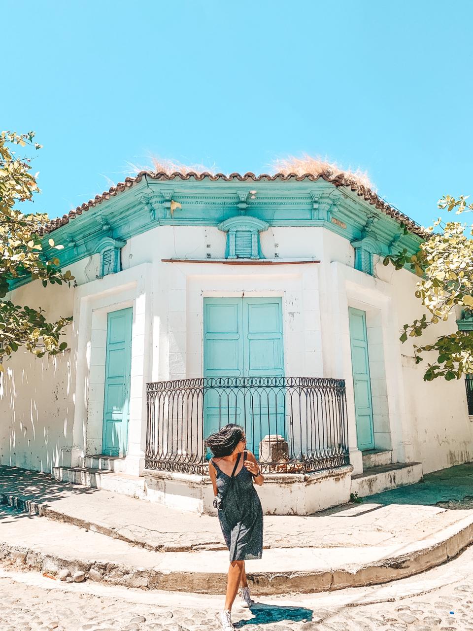 A historic building is showcased with a unique rounded structure featuring a light blue trim and double doors. A wrought iron railing surrounds a small terrace, while green trees provide a natural frame against a clear blue sky.