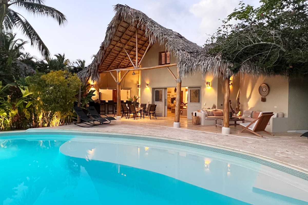 A tropical outdoor area features a large private pool with a curved edge. Rustic thatched-roof architecture provides shade over a dining area with seating. Surrounding lush greenery enhances privacy. A stone patio includes various lounge chairs for relaxation.