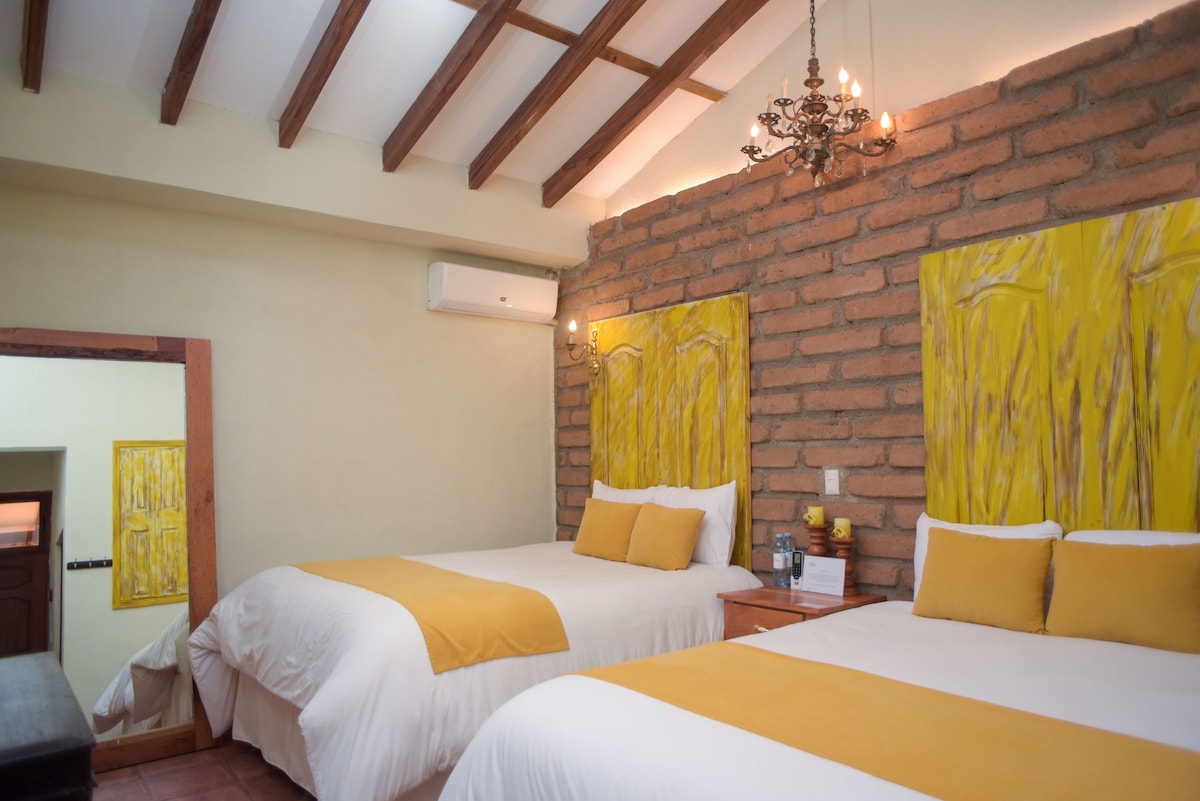 A inviting room features two spacious beds dressed in crisp white linens, accented by bright yellow pillows. Exposed wooden beams enhance the ceiling, while textured brick walls add character. A large mirror reflects the soft lighting from a decorative chandelier above.